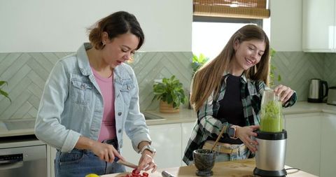 Mother and teen daughter preparing green smoothie, chopping strawberries in kitchen