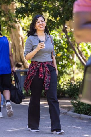 Smiling woman walking with to-go coffee on sunlit park path wearing casual plaid shirt