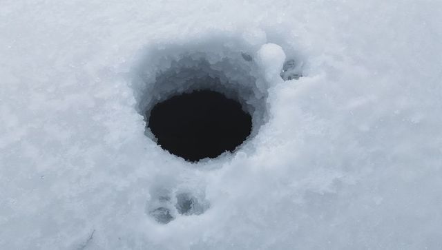 Showing circular dark ice hole with compacted rim and footprints on snow-covered surface