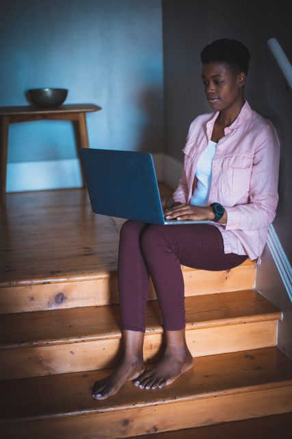 African american woman working at home sitting on stairs using laptop. staying at home in isolation during quarantine lockdown.