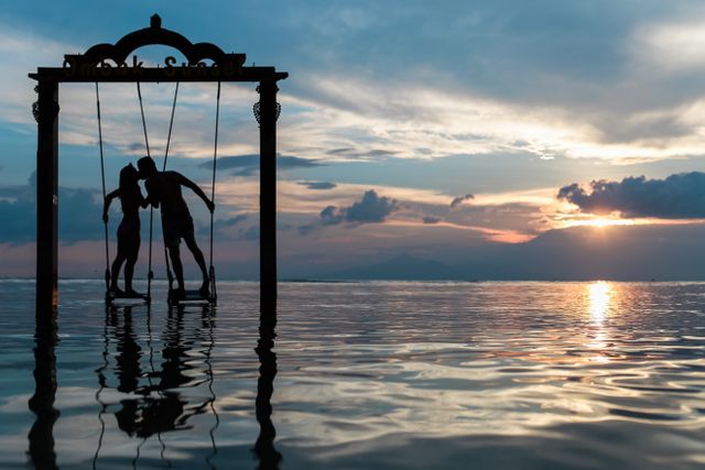 Romantic moment of couple on a swing during sunset, silhouetted by sky and water reflections. Suitable for themes of love, travel, romance, vacations.