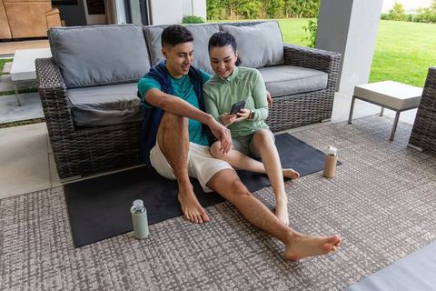 Couple relaxing on patio yoga mats sharing smartphone after outdoor workout session