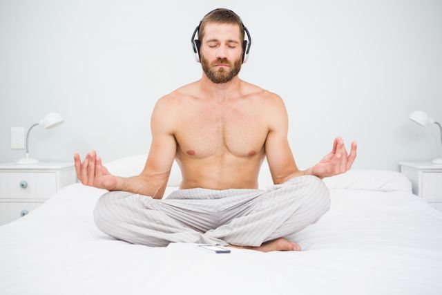 Man sitting cross-legged on bed, meditating with headphones on. Ideal for concepts of relaxation, mindfulness, and morning routines. Suitable for wellness blogs, mental health articles, and lifestyle websites.
