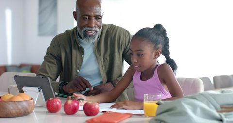 African american man tutoring young girl at kitchen table with tablet and homework