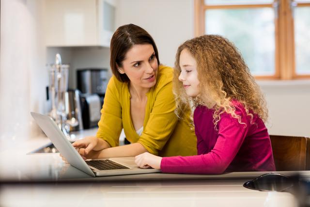 Mother and daughter are sitting at a kitchen counter, using a laptop together. The mother is wearing a yellow top, and the daughter is in a pink top with curly hair. They appear to be engaged in a conversation or learning activity. This image is ideal for use in family-oriented content, educational materials, technology advertisements, and lifestyle blogs.