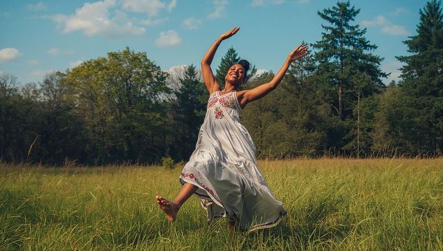 Joyful woman leaping through sunlit meadow in embroidered white flowy dress barefoot