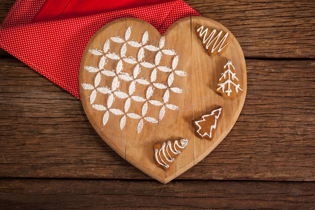 Heart-shaped wooden board with decorated gingerbread cookies, perfect for holiday-themed designs, Christmas cards, festive advertisements, and seasonal blog posts. The rustic wooden texture and red cloth add a warm, cozy feel suitable for winter and holiday promotions.