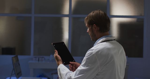 Male doctor using tablet in modern medical office at night. Ideal for illustrating telemedicine, modern healthcare technology, nocturnal activities in hospitals, or healthcare workers' night shifts. Useful for medical blogs, websites, advertisements, and articles related to healthcare IT, digital health solutions, or nighttime healthcare services.