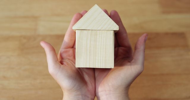Hands carefully holding a small wooden house model above a wooden table. Symbolizing protection, safety, and investment in real estate. Perfect for use in articles about property investment, real estate market, home ownership, or architectural models.