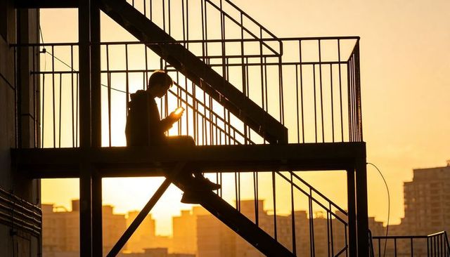 Silhouette person using smartphone on fire escape staircase at golden sunset urban skyline