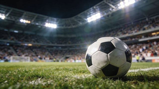 Scuffed soccer ball sitting on grass under stadium lights with packed crowd and goal