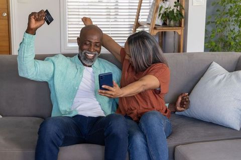 Senior couple celebrating online purchase using smartphone and credit card on sofa at home