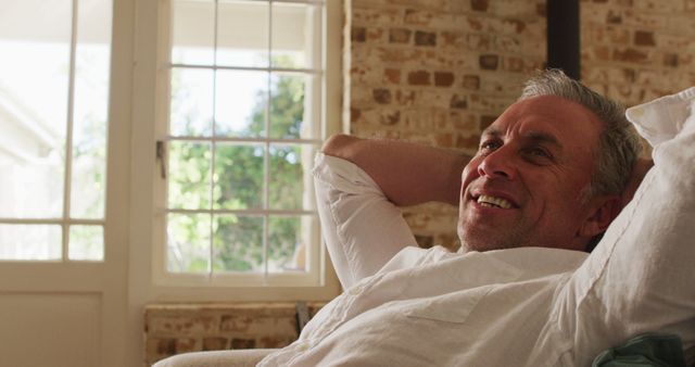 Middle-aged man reclining and smiling in bright, sunlit room with a brick wall backdrop. Perfect for use in lifestyle blogs, wellness articles, or advertising for home comfort products.