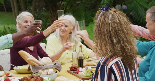 Image of diverse happy senior female and male friends eating lunch in garden, toasting. retirement lifestyle, spending quality time with friends.