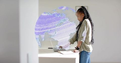 Standing woman interacting with holographic globe over laptop in minimalist workspace