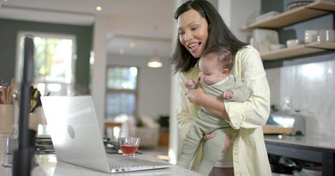 Mother engaging with work on laptop while holding baby in cozy kitchen
