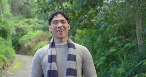 Asian man smiling on forest trail wearing knit sweater and striped scarf, enjoying nature