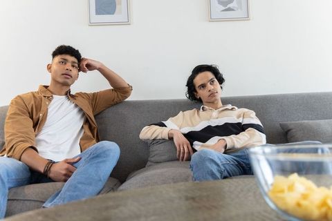 Diverse young men sitting on gray sofa looking pensive while watching tv in living room