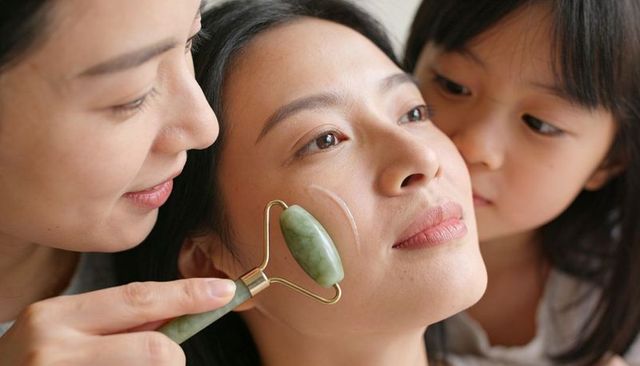 Asian woman receiving jade roller facial from caregiver with child observing skincare