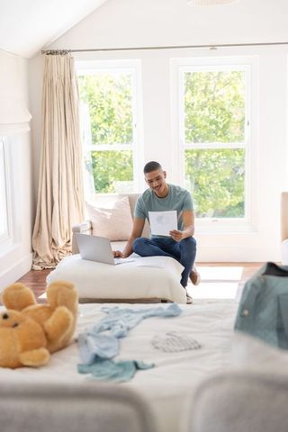 Young man working remotely in sunlit bedroom with laptop, printed charts and cozy decor