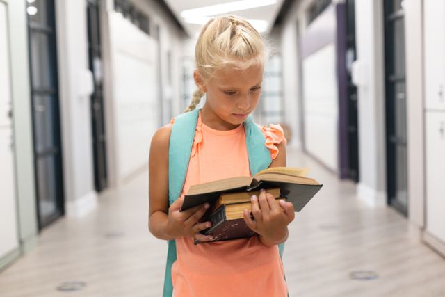 Young Caucasian schoolgirl standing in a school corridor, reading a book with concentration. She is wearing a backpack and appears to be deeply engaged in her reading. This image is ideal for educational materials, school promotions, childhood learning resources, and articles about student life.