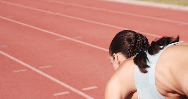 This image depicts a female sprinter crouching and preparing at the starting line of a running track. It captures the athlete's readiness and focus before a race. Ideal for use in sports advertisements, fitness promotions, articles on athletic training, or motivational materials targeting aspiring athletes.