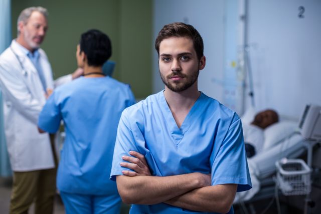 Male nurse standing with arms crossed in a hospital ward, exuding confidence and professionalism. In the background, other medical staff are engaged in conversation, and a patient is resting in a bed. This image can be used for healthcare-related content, medical websites, hospital brochures, and articles about nursing and patient care.