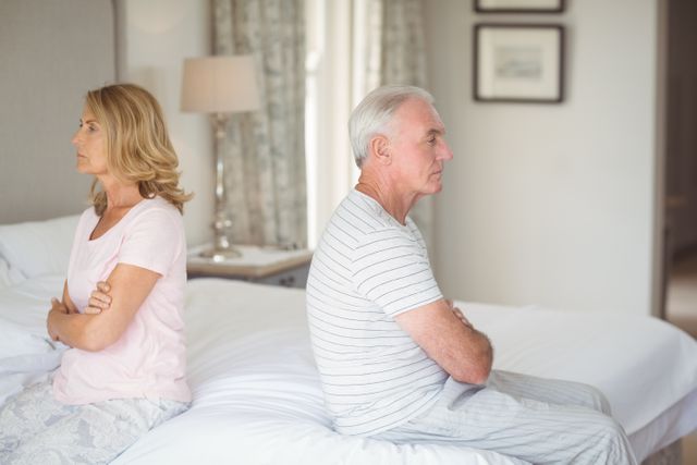 This image depicts an elderly couple sitting back to back on a bed, both appearing upset and distant. It can be used to illustrate themes of relationship issues, conflict resolution, emotional tension, and challenges in mature relationships. Suitable for articles, blogs, or advertisements related to marriage counseling, senior relationships, and emotional well-being.