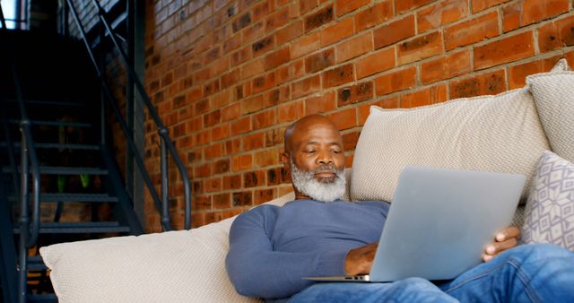 An older man with a beard uses a laptop while sitting on a comfortable couch. The setting includes a brick wall in the background, creating a cozy and warm atmosphere. Useful for depictions of remote work, casual business activities, or leisure time at home.