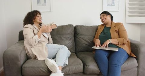 Two friends chatting on gray sofa wearing blazers and jeans, cozy casual home conversation