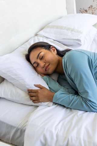 Asian woman sleeping peacefully hugging white pillow wearing light blue loungewear in cozy bedroom
