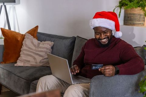 African american man wearing santa hat shopping online on laptop holding credit card