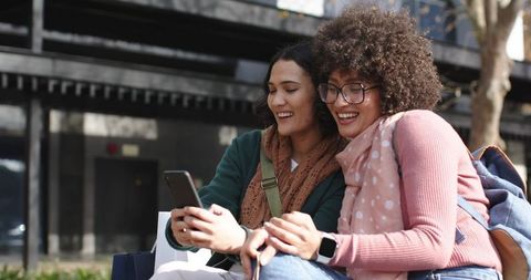 Two women sharing smartphone and shopping bags while sitting on urban bench, smiling