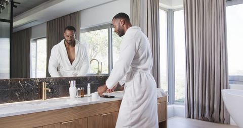 Mature man in white bathrobe arranging toiletries on marble vanity in bright bathroom daylight