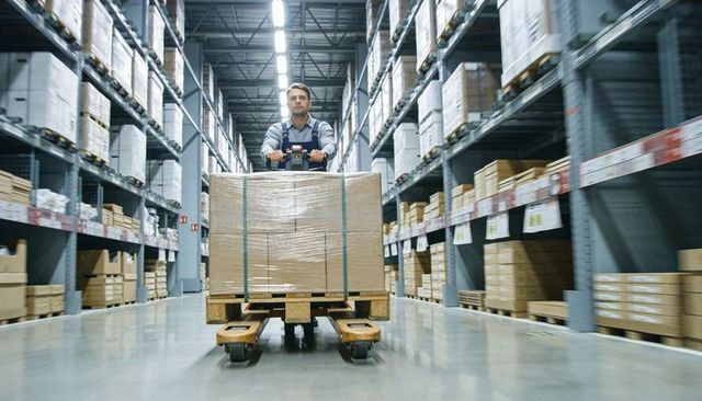 Worker operating pallet jack with shrink-wrapped pallet in large warehouse aisle