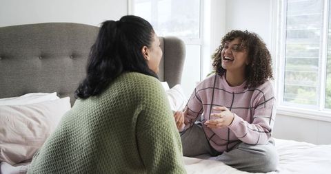 Two diverse women laughing and chatting on bed wearing cozy sweaters in bright bedroom