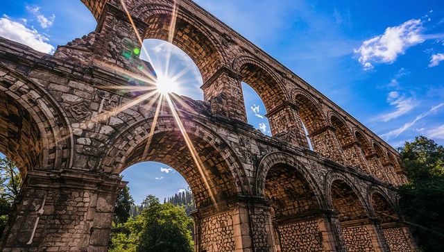 Sunburst framing towering roman aqueduct rising over wooded valley with arched spans
