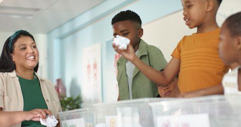 African american teacher smiling while guiding children sorting paper into clear bins