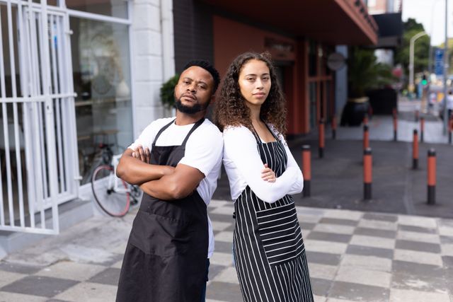 Two cafe coworkers, one man and one woman, standing back to back with arms crossed on an urban sidewalk. Both wear aprons, indicating their roles in the cafe. This image is ideal for illustrating concepts of teamwork, small business, and the hospitality industry. It can be used in marketing materials, websites, or articles related to cafe culture, diversity in the workplace, and entrepreneurship.