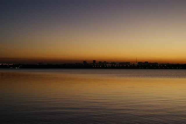 Depicting a tranquil scene of a sunset over calm waters with a city skyline reflecting in the foreground. Suitable for use in peaceful and serene designs, travel websites, urban promotions, and nature-themed projects. Also ideal for relaxation-themed content or for illustrating evening cityscapes.