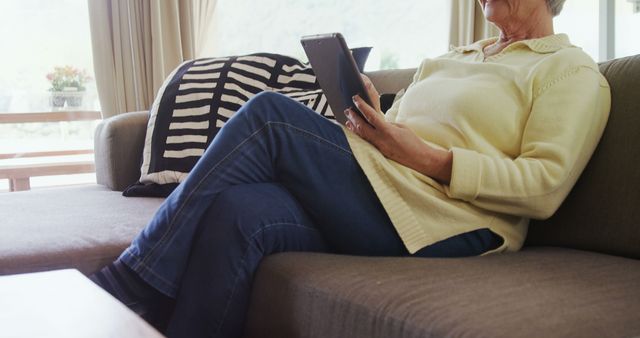 Senior woman lounging on a couch at home, utilizing a tablet for leisure or information. Suitable for articles or advertisements on senior lifestyle, technology use among the elderly, or comfortable home settings. Also ideal for content focused on digital literacy and the importance of technology in maintaining connections and entertainment for seniors.