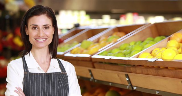 Woman working in a grocery store standing in front of a fruit display, smiling. She wears an apron, indicative of her role in providing customer service. Ideal for use in content about grocery stores, customer service, retail industry, fresh produce, and healthy eating. This image conveys friendliness and professionalism in a retail or supermarket setting.