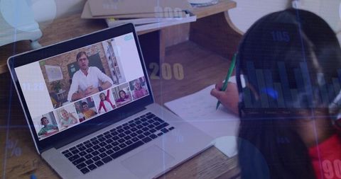 School-age student taking notes during online class at home desk with laptop, headphones