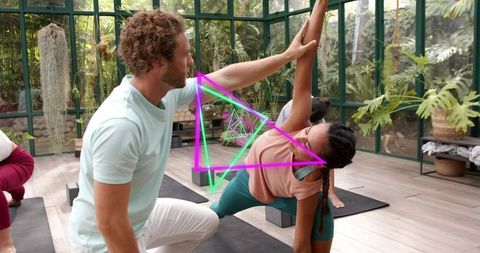 Yoga instructor assisting student with side angle pose inside glass greenhouse with plants
