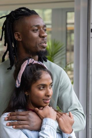 Interracial couple embracing near window in light-filled interior, intimate lifestyle moment
