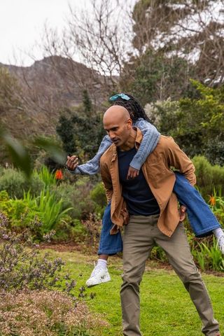 African american father carrying daughter piggyback walking through garden bonding outdoors