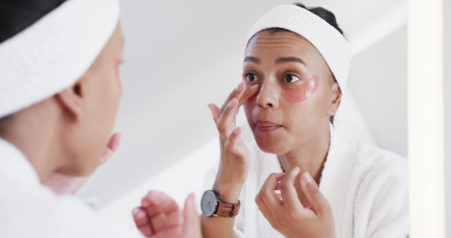 Woman applying eye patches while looking in bathroom mirror, wearing white bathrobe and headband. Ideal for articles about beauty routines, skincare tips, self-care practices, or wellness lifestyle promotions.
