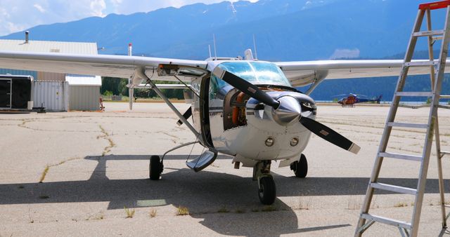 Small private airplane parked on a runway in front of scenic mountain range and clear sky. Propeller plane resting on tarmac near hangar and ladder. Ideal for use in articles about private aviation, travel, and outdoor adventure.