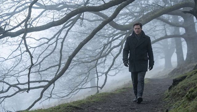 Solitary man walking misty woodland path under bare branches wearing dark coat and scarf