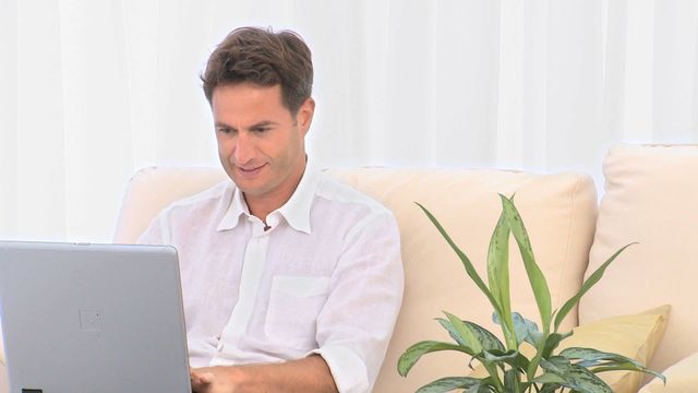 Man sitting on sofa, using laptop with a smile. Background includes a white curtain and a plant. Suitable for use in articles about remote work, home comfort, or technology use at home.